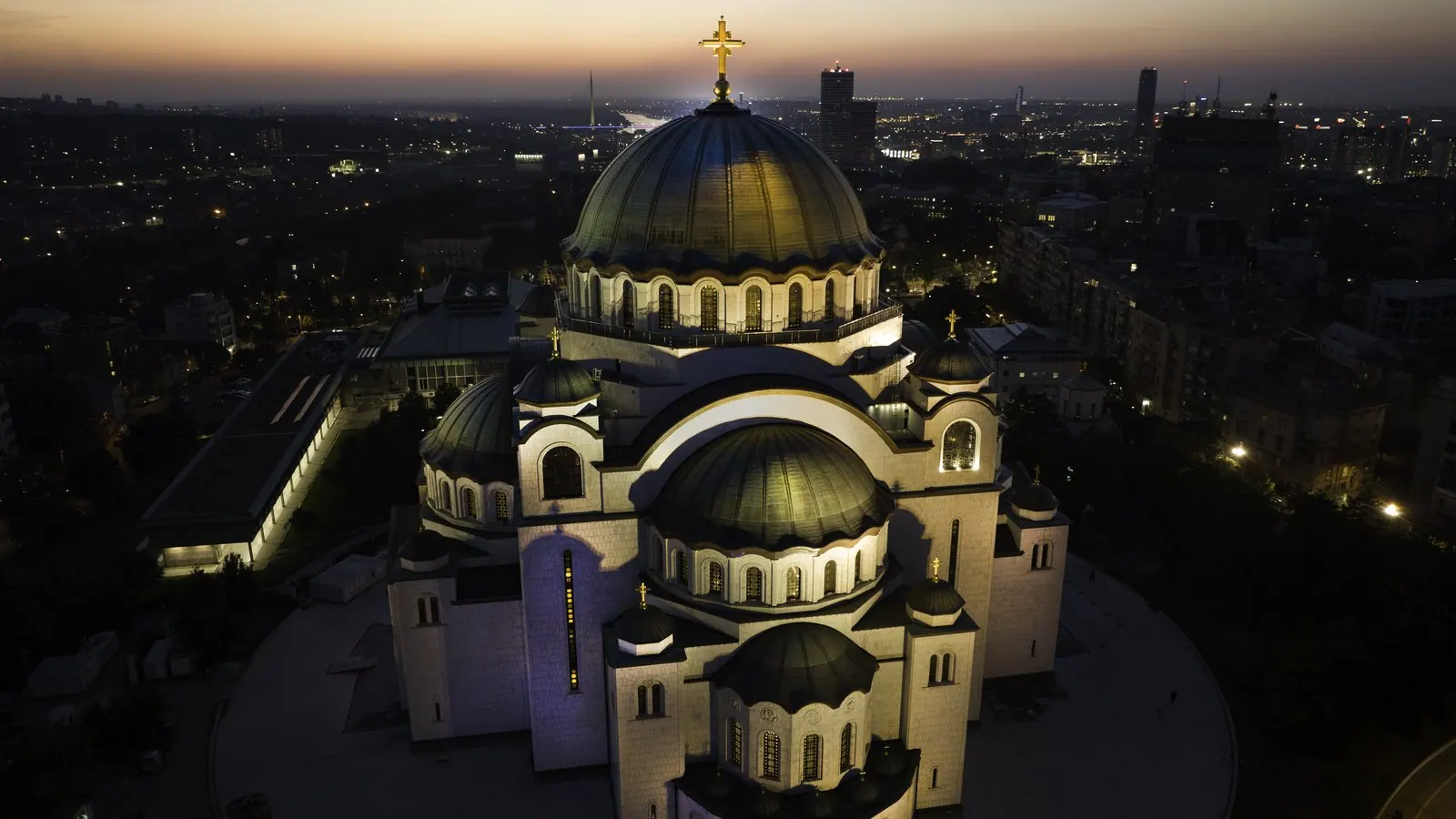 Luftaufnahme der Kirche des heiligen Sava in Belgrad, Serbien, in der Dämmerung, mit angestrahlten weißen Kuppeln und vergoldetem Kreuz über den Lichtern der Stadt.