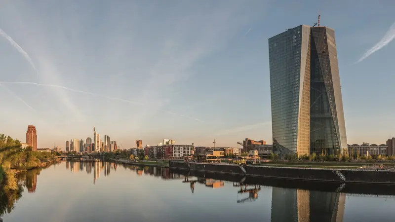 Der gläserne Turm der Europäischen Zentralbank am Main in Frankfurt im Morgenlicht, mit der Skyline der Stadt und ihrer Spiegelung im Wasser.