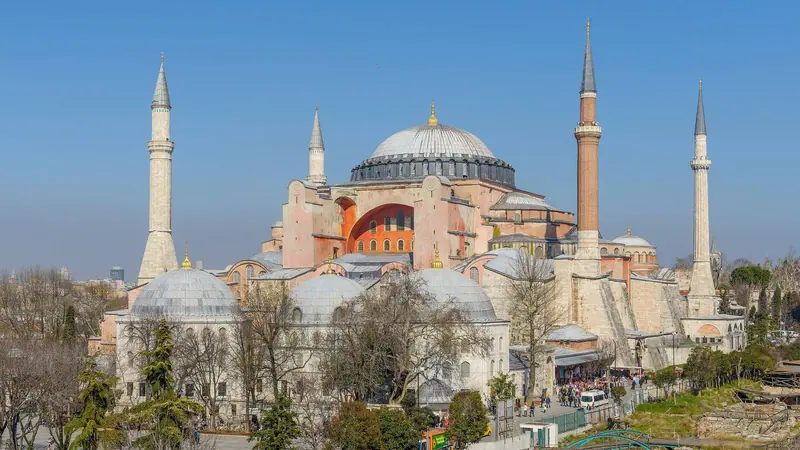 Die Hagia Sophia in Istanbul mit großer Kuppel, Strebepfeilern und vier Minaretten unter klarem blauem Himmel, kahle Bäume im Vordergrund.