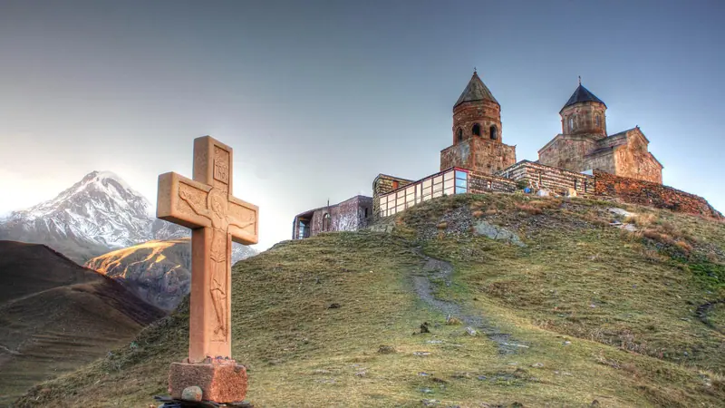 Die Dreifaltigkeitskirche von Gergeti auf einem grünen Bergrücken unter dem verschneiten Kasbek in Georgien, vorne ein steinernes Kreuz.