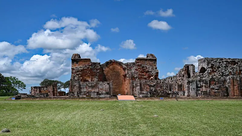 Die Ruinen der roten Sandsteinkirche der Jesuitenmission La Santísima Trinidad de Paraná in Paraguay unter blauem Himmel mit weißen Wolken.