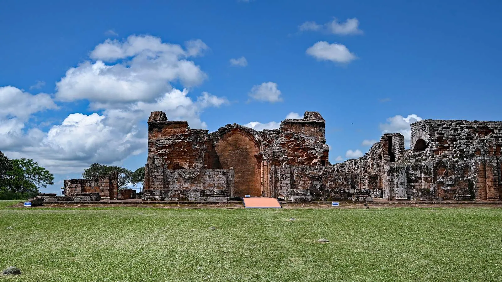 Die Ruinen der roten Sandsteinkirche der Jesuitenmission La Santísima Trinidad de Paraná in Paraguay unter blauem Himmel mit weißen Wolken.