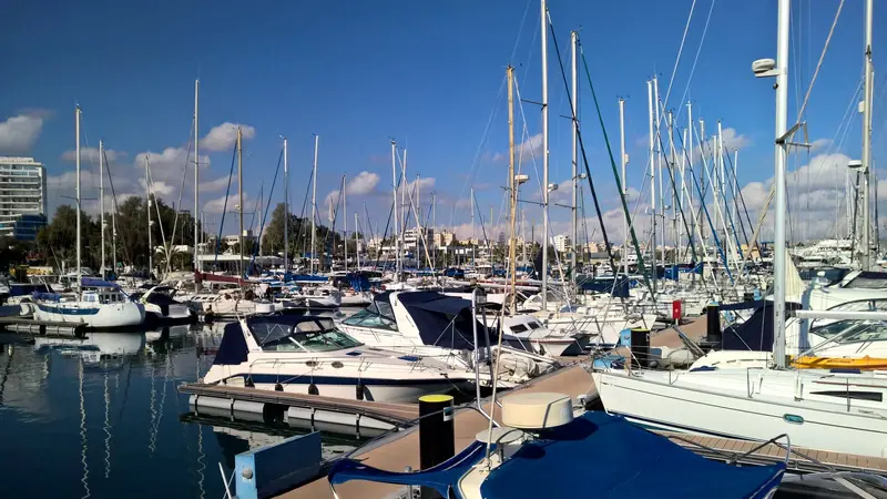 Segelboote und Motoryachten in der Marina von Larnaka, Zypern, an einem sonnigen Tag, dahinter die Gebäude der Uferpromenade unter blauem Himmel.